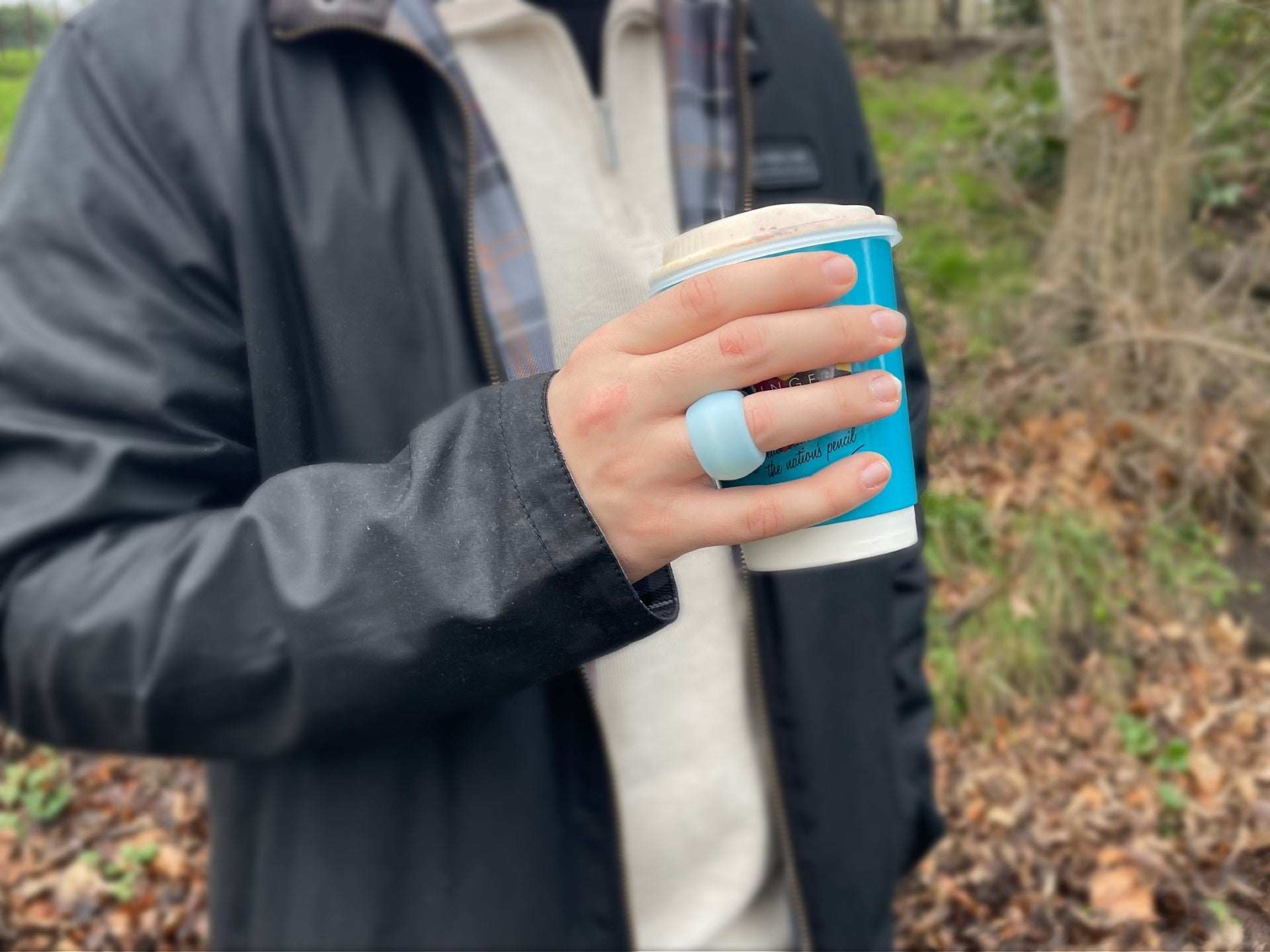 A man wears a blue ringguard ring protector on his hand to cover jewellery. He holds a coffee cup. Woodland background.